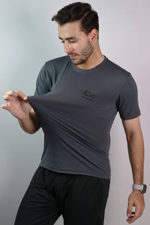 A man wearing a dark gray athletic t-shirt with a black logo on the chest, stretching the fabric to demonstrate its elasticity. He is also wearing black athletic pants against a plain light gray studio background.