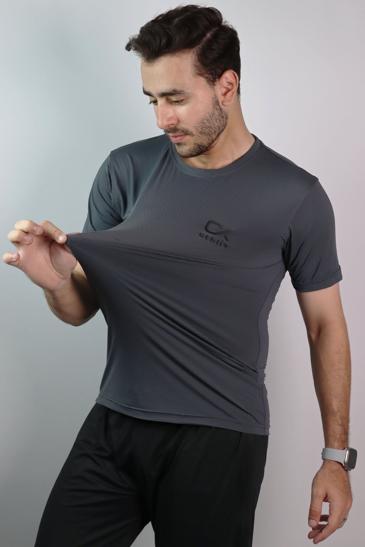 A man wearing a dark gray athletic t-shirt with a black logo on the chest, stretching the fabric to demonstrate its elasticity. He is also wearing black athletic pants against a plain light gray studio background.