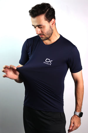 A man wearing a navy blue athletic t-shirt with a white logo on the left chest, stretching the fabric to show its elasticity. The background is a plain light gray studio backdrop.
