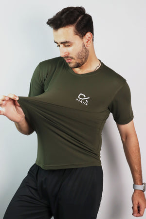 A man wearing an olive green athletic t-shirt with a white logo on the left chest, stretching the fabric to show its elasticity. The background is a plain light gray studio wall.