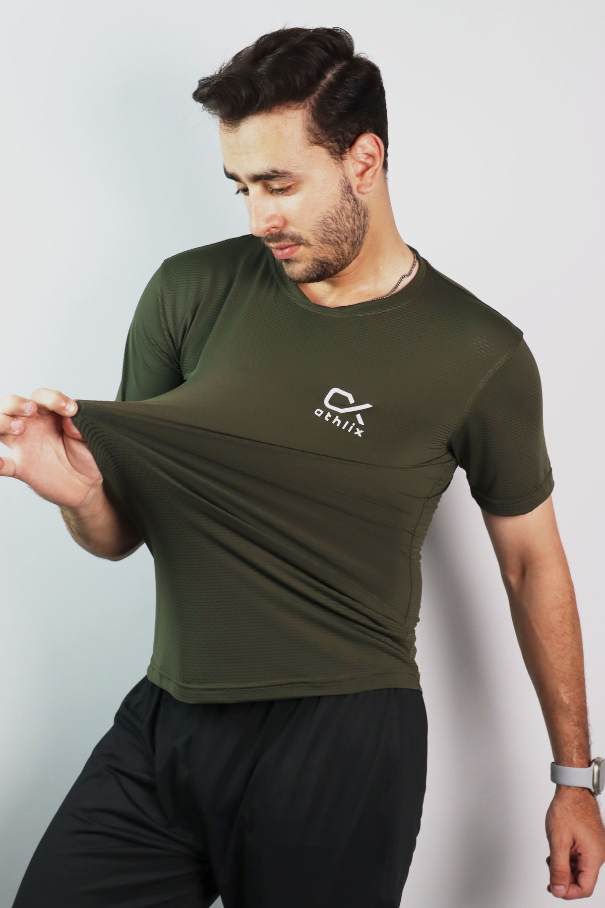 A man wearing an olive green athletic t-shirt with a white logo on the left chest, stretching the fabric to show its elasticity. The background is a plain light gray studio wall.